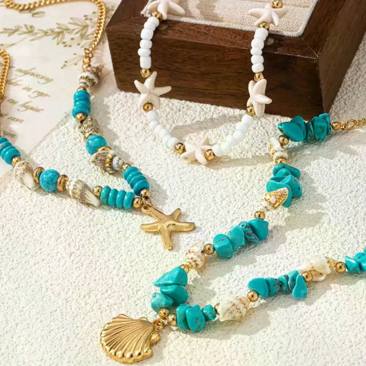 A close-up of layered beaded necklaces with turquoise, white, and gold elements, including starfish and shell charms, arranged on a textured white surface with a wooden box in the background | DIVINE TIMES by Shopp Java