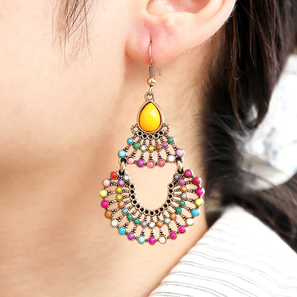 A close-up of a person's ear wearing a large, dangling statement earring. The earring features a gold-toned metal frame with a bright yellow teardrop stone at the top and a wide, scalloped lower section adorned with many small, colorful rhinestones in pink, blue, green, orange, and purple | DIVINE TIMES by Shopp Java 