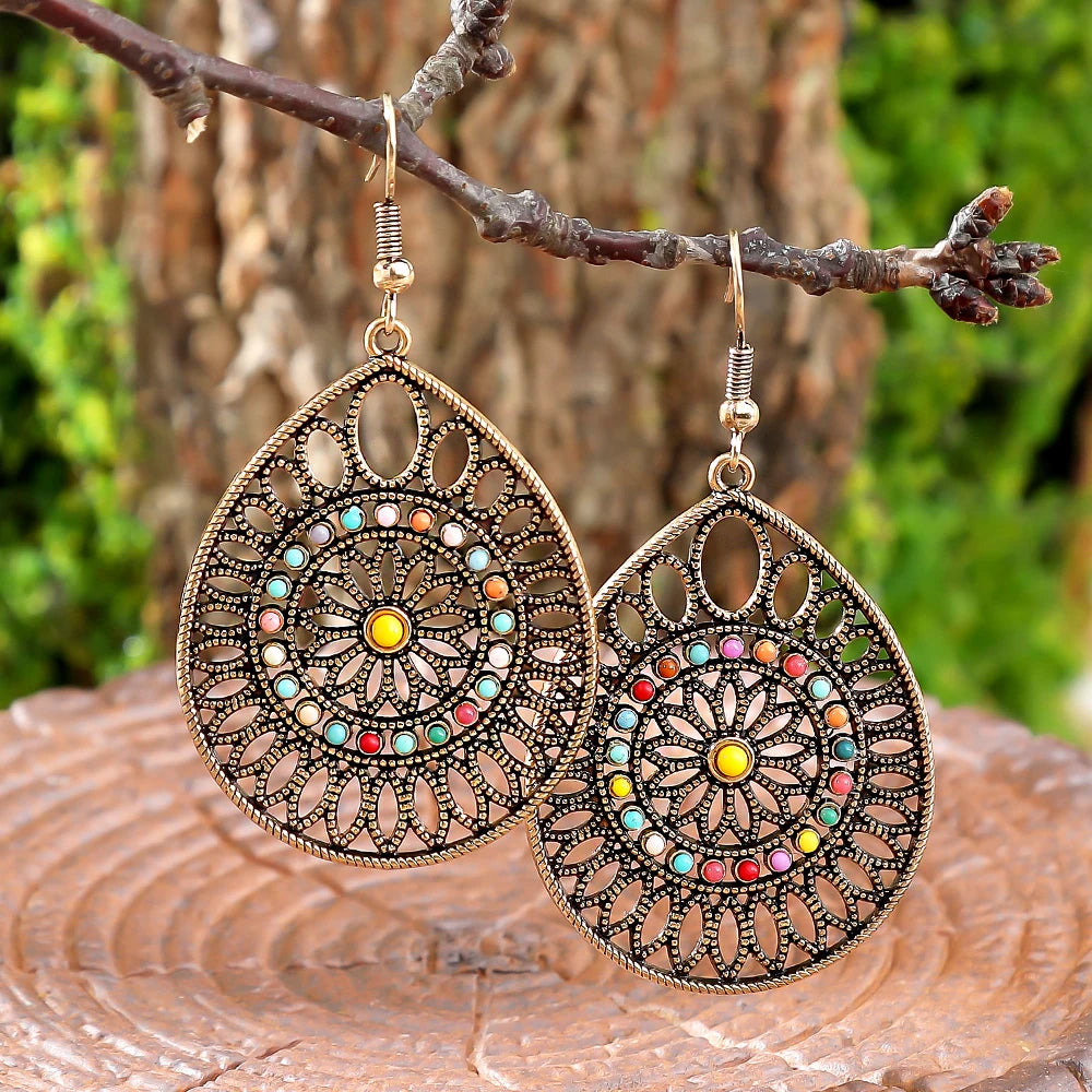 A pair of large teardrop-shaped dangling earrings with ornate filigree cutouts and a circular multicolor gemstone pattern, hanging from a tree branch with a blurred forest background | DIVINE TIMES by Shopp Java 