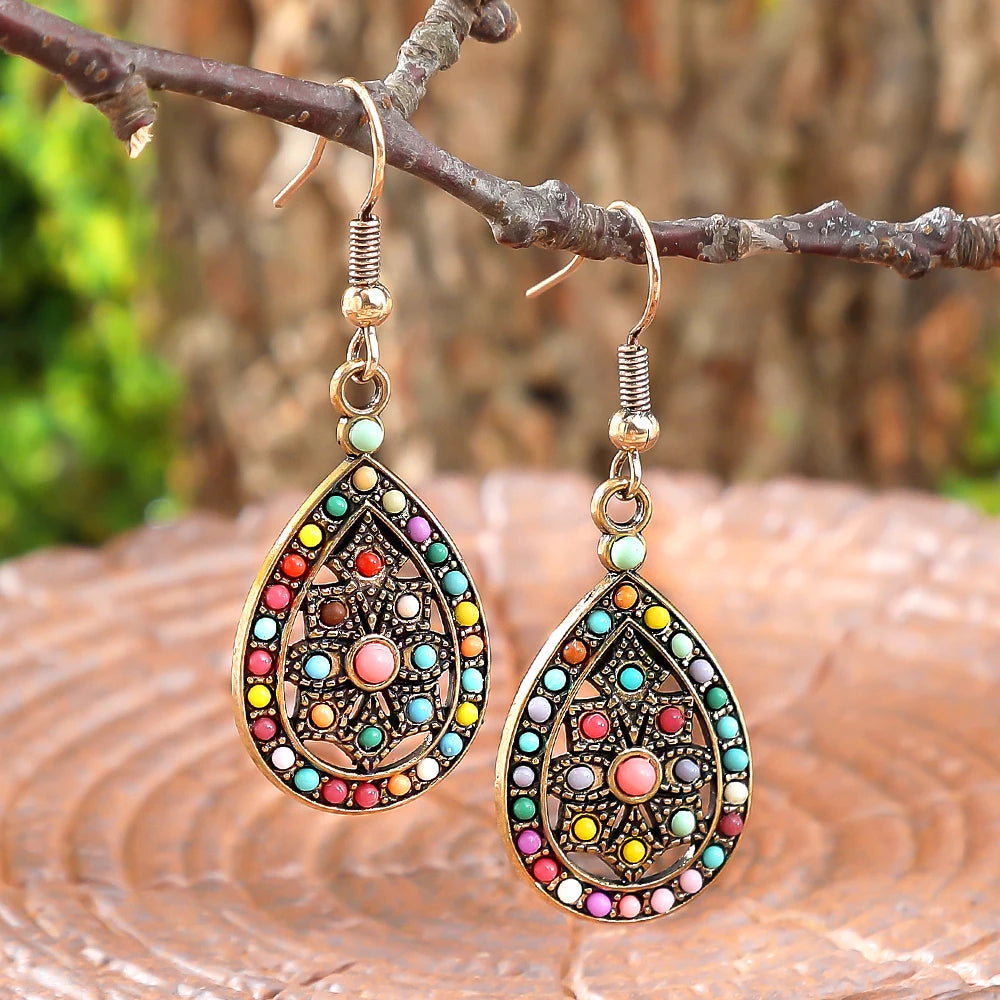 A pair of ornate teardrop-shaped dangling earrings with colorful bead accents and a central pink stone, hanging from a tree branch over a wooden surface with a blurred natural background. | DIVINE TIMES by Shopp Java 