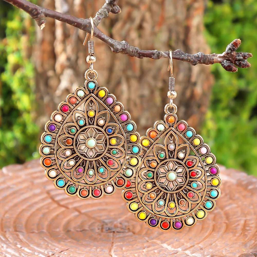 A pair of ornate gold-toned dangle earrings with colorful gemstone accents, hanging from a tree branch outdoors with blurred green foliage and a wooden surface in the background | DIVINE TIMES by Shopp Java 