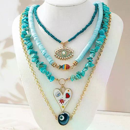 A multi-layered bohemian-style necklace set displayed on a white mannequin bust. The set features a turquoise beaded chain, a light blue cord chain, a turquoise stone chip chain, a gold chain with a white heart pendant decorated with colorful floral motifs and a dark blue evil eye charm, and a turquoise evil eye bead pendant | DIVINE TIMES by Shopp Java